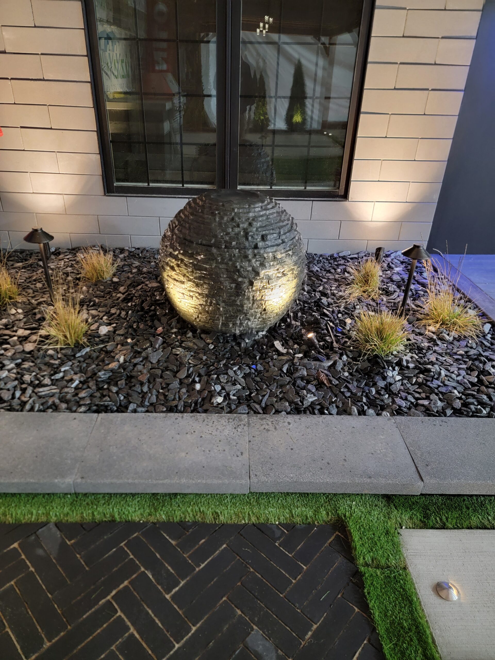 Round textured stone fountain in a landscaped rock bed beside a tiled wall