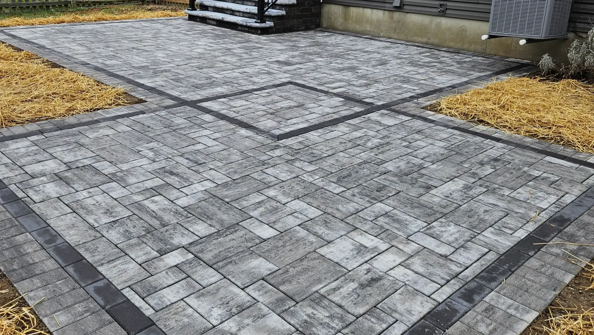 Gray interlocking paver patio with dark edging beside a house and landscaped beds